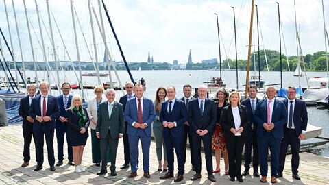 Ein Gruppenfoto mit rund 15 Personen in geschäftlicher Kleidung (Anzüge und Kostüme), die nebeneinander an einer Hafenpromenade vor einer Kulisse aus zahlreichen Segelbooten und einer Stadt-Silhouette im Hintergrund posieren.