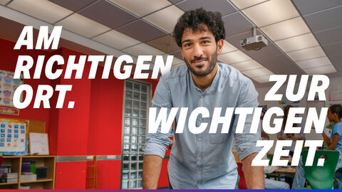 Ein Sozialpädagoge steht im Klassenzimmer. Text auf dem Bild: Am richtigen Ort. Zur wichtigen Zeit.