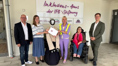 Mehrere Menschen stehen mit einem Roboter vor einer Wand und lachen in die Kamera. Eine Person sitzt im Rollstuhl